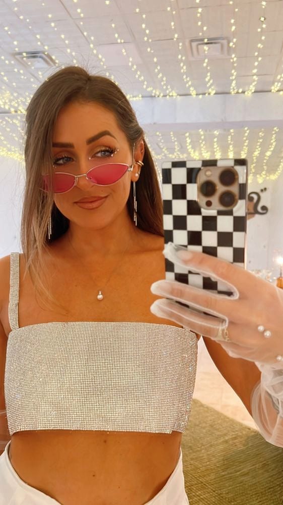 Mirror selfie of a woman in sunglasses and a sparkly top, string lights behind her